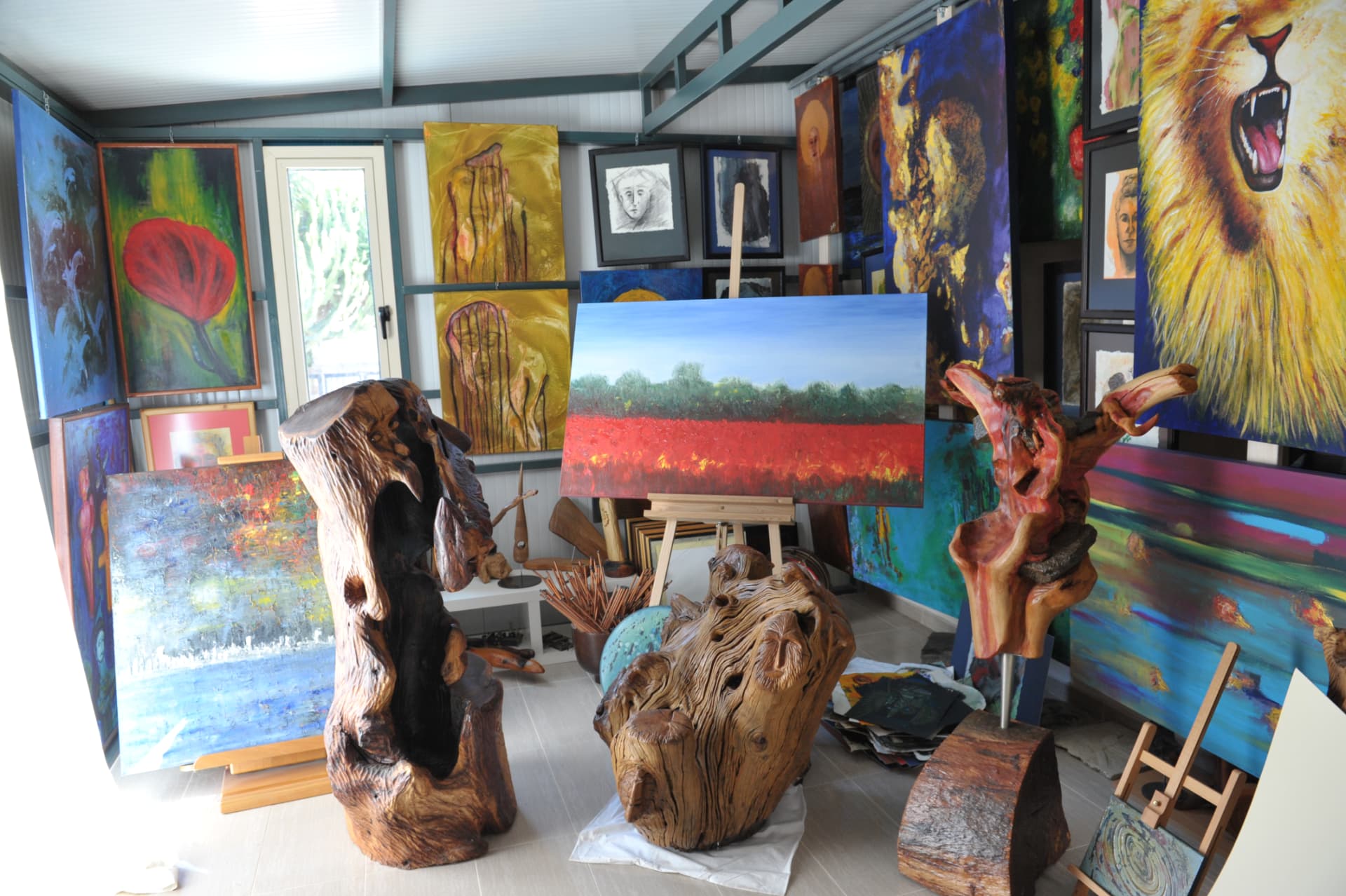 Studio interior with three carved wood sculptures in the foreground, a landscape painting on a wooden tripod easel, and walls covered with colourful canvases